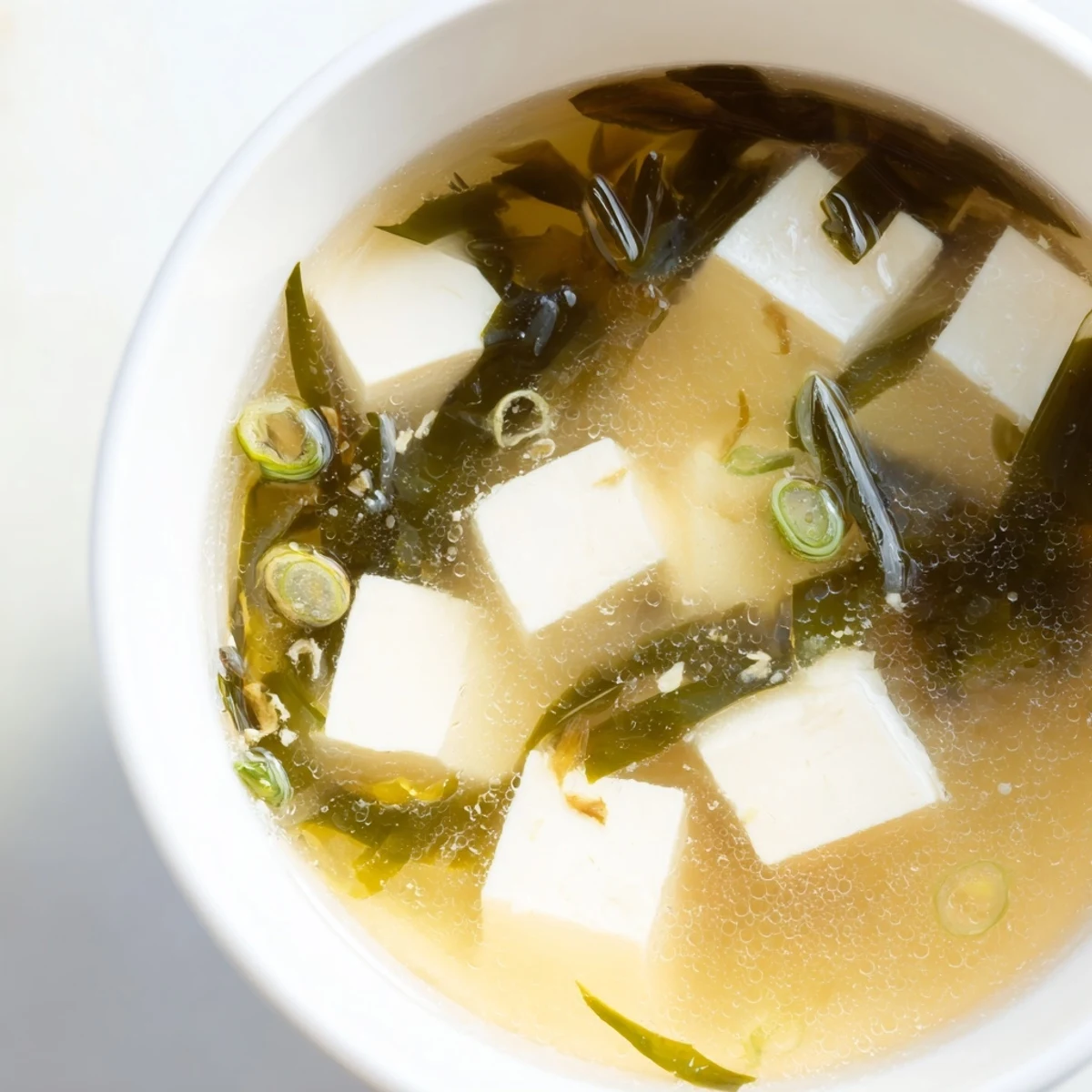 Bol de soupe miso avec tofu soyeux, wakame tendre et ciboulette fraîche, présenté sur fond clair.
